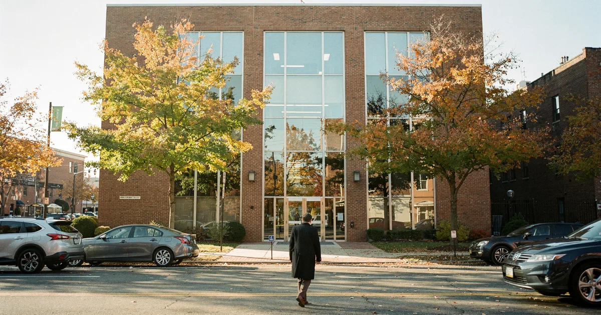 A person walks toward a brick and glass office building on a tree-lined street on an autumn morning