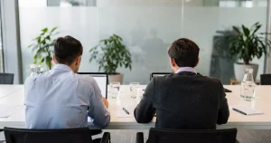 Two analysts work side by side at laptops in a glass-walled conference room